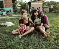 Mujeres y una niña sentadas sobre el césped, en un entorno rural, con viviendas de ladrillos al fondo. Comunidad Eyiyoquibo
