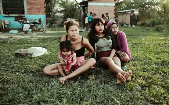 Mujeres y una niña sentadas sobre el césped, en un entorno rural, con viviendas de ladrillos al fondo. Comunidad Eyiyoquibo